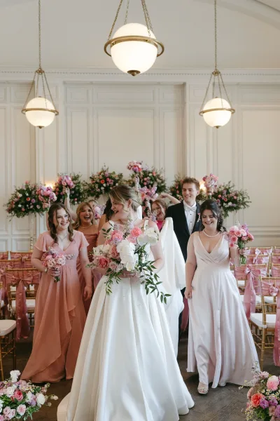 Bride with wedding party walking down the aisle, holding pink and white bouquet, in a white-paneled indoor ceremony room with chandeliers