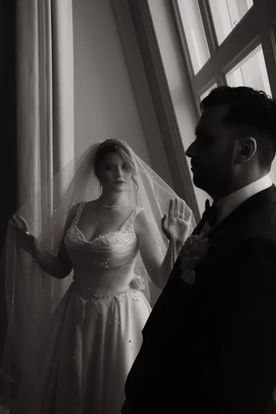 Couple portrait in black and white wedding portrait style, bride in pearl veil and dress with groom in tuxedo by tall window curtains