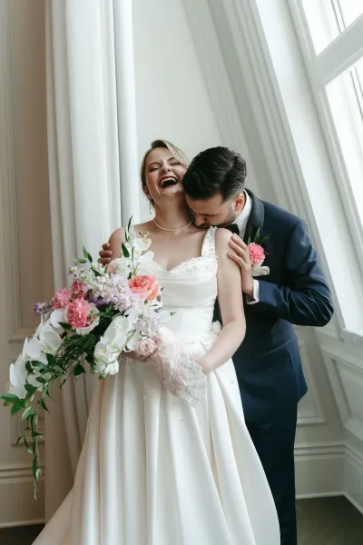 Couple portrait of bride and groom embrace by a large window, groom kissing her shoulder as she laughs holding a pink bouquet
