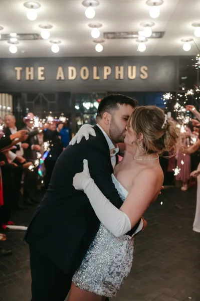 Wedding kiss during a sparkler send off, bride in strapless sequin mini dress and long white gloves with groom in tuxedo under string lights outside hotel entrance at night