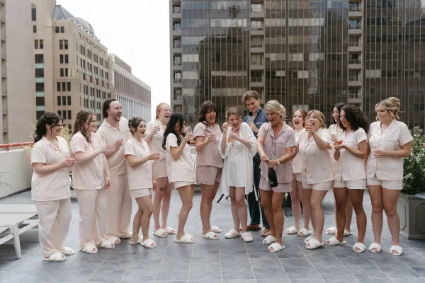 Bridal party getting ready as bridesmaids matching pajamas raise champagne flutes on a rooftop terrace with city buildings behind them