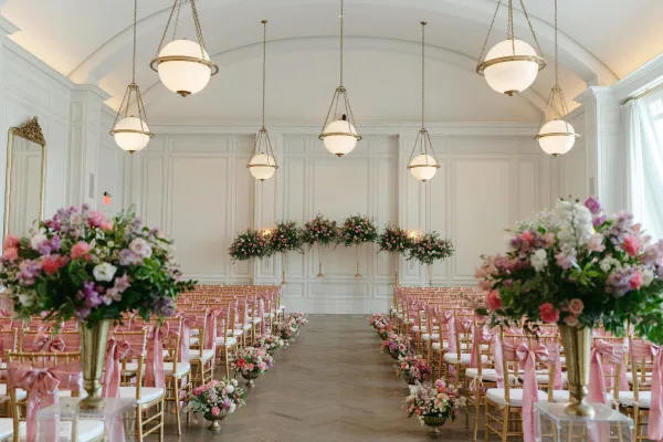 Ceremony setup with gold chiavari chairs and pink bows lining a wedding ceremony aisle, framed by floral urns under globe chandeliers