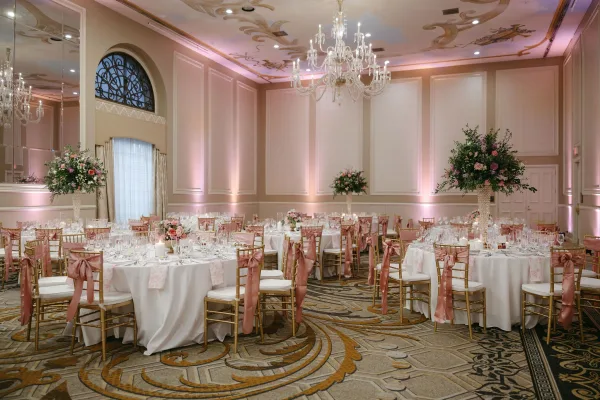 Reception tablescape with round wedding tables, blush runners, gold Chiavari chairs, and tall florals under a crystal chandelier in a ballroom