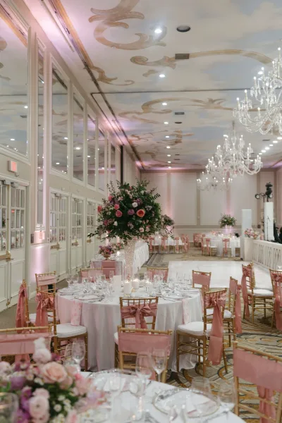 Reception tablescape with round wedding tables, white linens, tall floral centerpiece in a crystal vase, candles, and gold chiavari chairs in an ornate ballroom