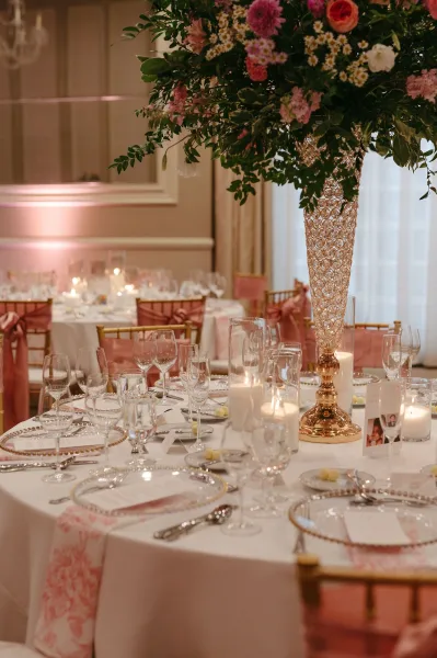 Reception tablescape with tall floral centerpiece in a crystal vase, candlelit place settings, gold Chiavari chairs, and pink sashes in a ballroom
