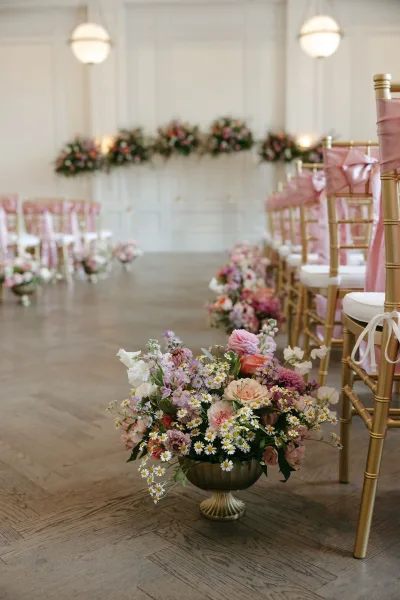 Ceremony aisle decor with wedding aisle flowers in gold compote vases, lined by pink-sashed chiavari chairs on a herringbone wood floor.