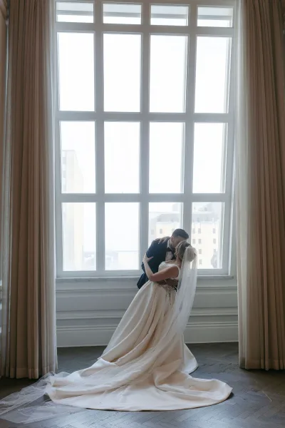Wedding kiss portrait of groom dipping bride, her long veil train flowing on a wood floor by a large window with city buildings beyond