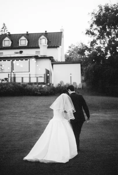 Couple portrait of a bride and groom walking across a grass lawn at dusk, with warm string lights twinkling by a house and trees.