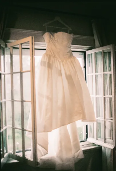 Wedding dress, the strapless wedding dress, hangs on a hanger in an open window with curtains framing outdoor greenery