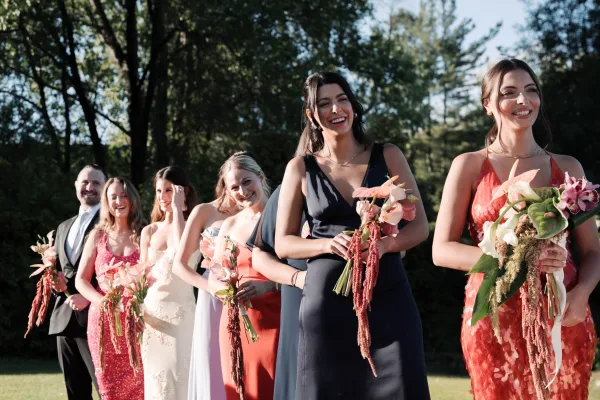 Wedding party portrait featuring a cheerful bridesmaids photo lineup, bouquets in hand, on a garden lawn framed by trees and blue sky.