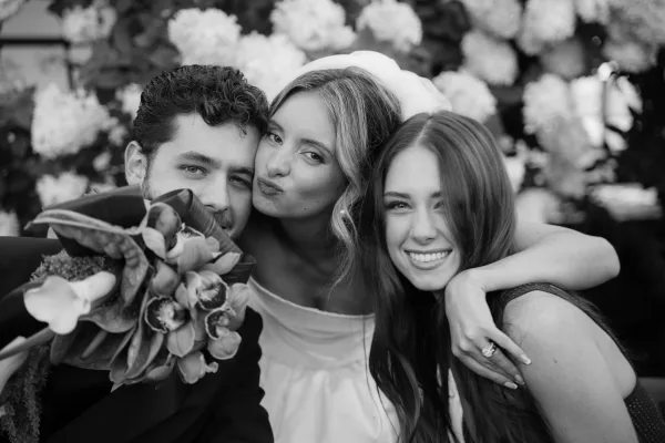 Wedding group portrait of a bride with friend and groom cheek to cheek, her bouquet framed by flowering shrubs and lush garden greenery.