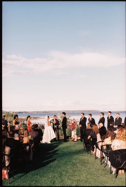 Ceremony moment at an outdoor wedding ceremony as the couple stands by vibrant altar flowers on a lakeside lawn beneath open sky and clouds.