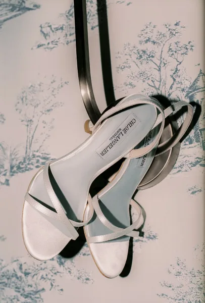 Bridal shoes, strappy wedding sandals with satin finish, hang from a metal hook against toile wallpaper in a delicate pattern.