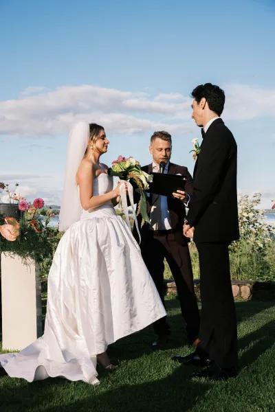 Ceremony moment as a couple exchanging vows beside calm water under a blue sky, the bride holding a bridal bouquet as they face each other.