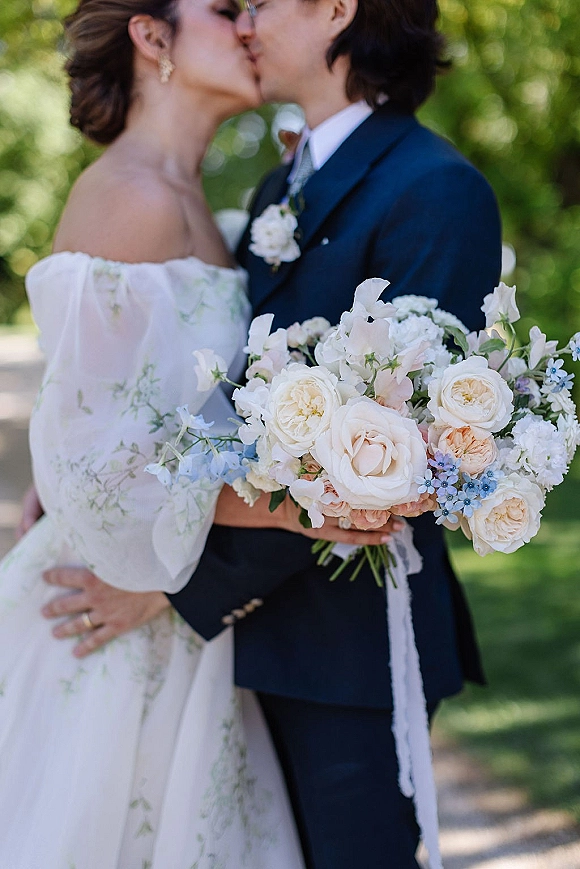 Wedding kiss as bride and groom kiss beside a white rose bouquet in a sunlit garden, groom’s boutonniere and her gown visible