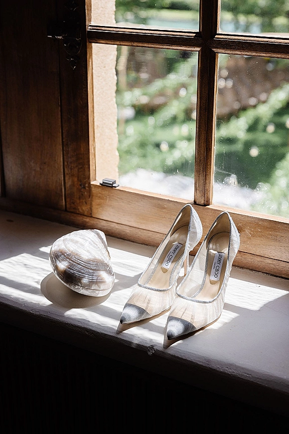 Wedding shoes and bridal heels with sheer mesh and metallic toe caps beside a shell clutch on a sunlit windowsill with greenery outside