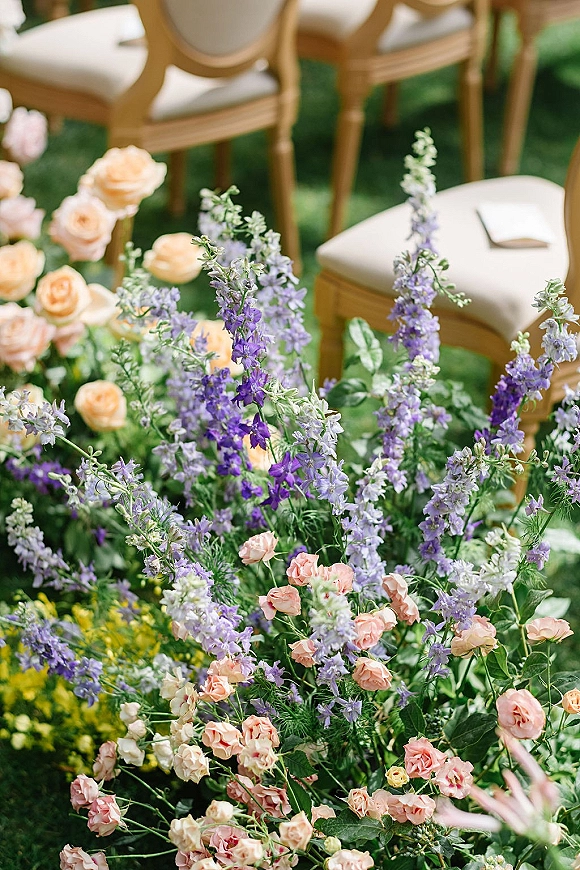 Ceremony aisle flowers in a garden ceremony aisle flowers display of blush roses, purple delphinium and greenery beside upholstered chairs on grass lawn
