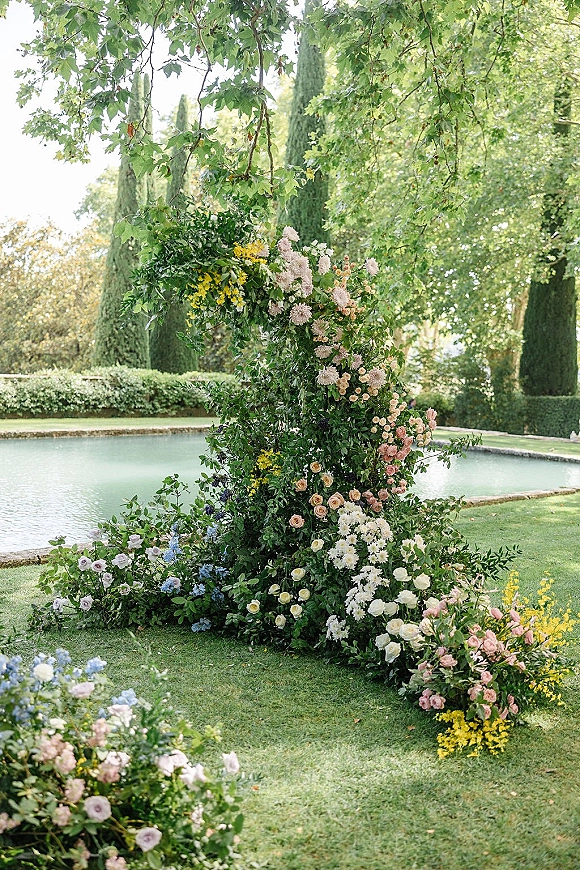 Wedding ceremony arch with asymmetrical floral arch of pink and cream roses, dahlias, and greenery on a garden lawn by a pond