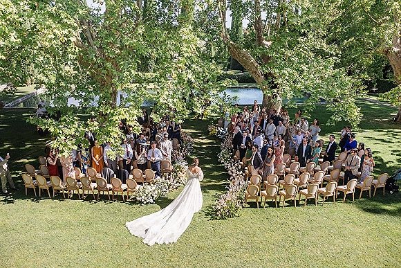 Ceremony processional as bride walking down aisle from behind, long train gown trailing past floral-lined chairs on garden lawn by pond