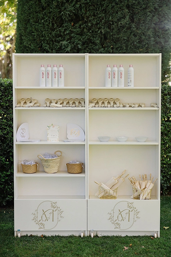 Wedding welcome shelf with water bottles, handheld fans, programs, and parasols in woven baskets on a lawn by hedges and evergreens