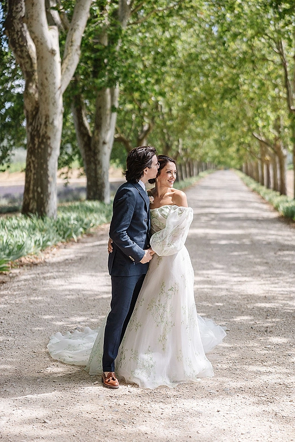 Couple portrait of groom kissing bride’s cheek as they hug on a tree-lined gravel road, her strapless floral gown with tulle train in sun