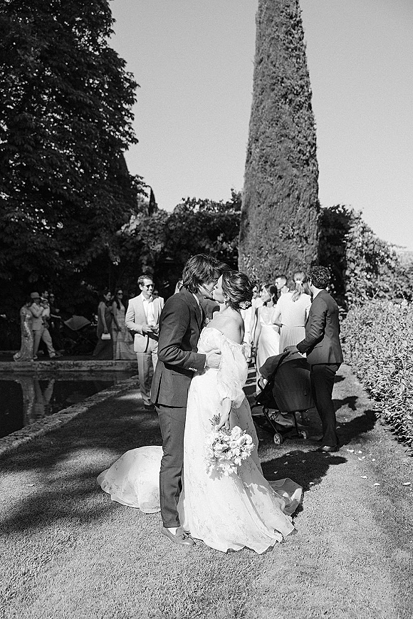 Wedding kiss as the bride and groom kissing, she holds a bouquet in a veil while guests watch on a garden lawn by a pond