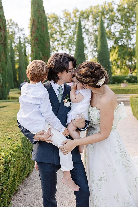 Wedding family portrait of bride and groom with kids, parents kissing while holding a baby on a gravel path beside garden hedges