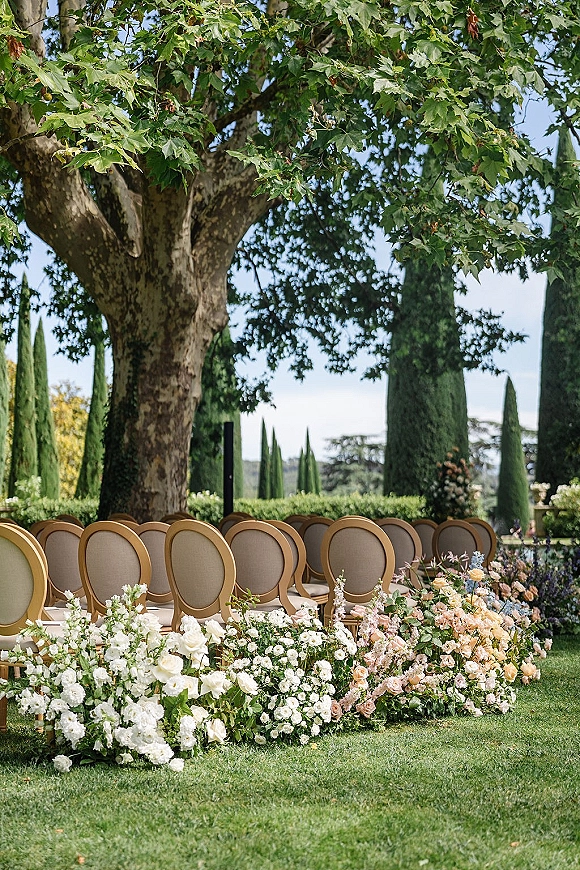 Ceremony seating with outdoor wedding ceremony chairs in rows, lined with white and blush rose and hydrangea ground florals on a lawn beneath trees