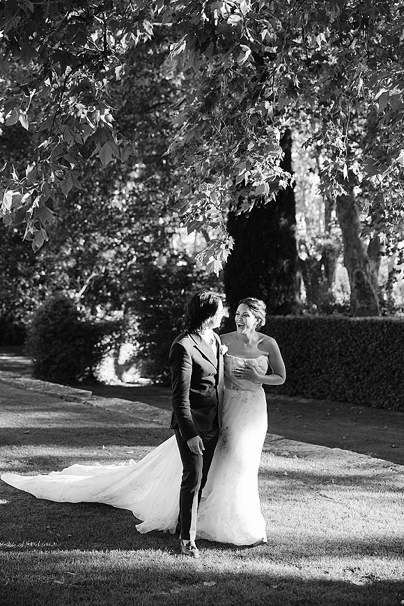 Couple portrait in black and white, bride in strapless gown with long train and groom in suit laughing under sunlit garden trees