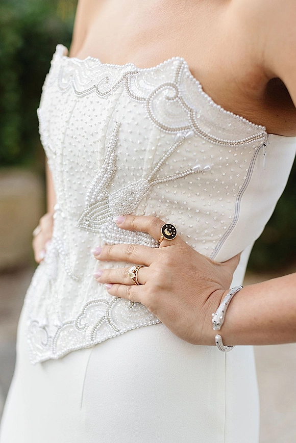 Wedding dress close-up showing a beaded wedding dress bodice with pearl beading, bride’s hand on waist with ring stack on an outdoor walkway