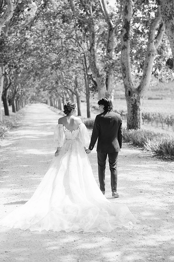 Wedding couple portrait of bride and groom walking away holding hands, her long gown train and updo on a sunlit tree-lined road