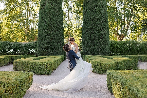 Couple portrait of groom lifting bride in a strapless gown with long train on a gravel path amid trimmed hedges and cypress trees