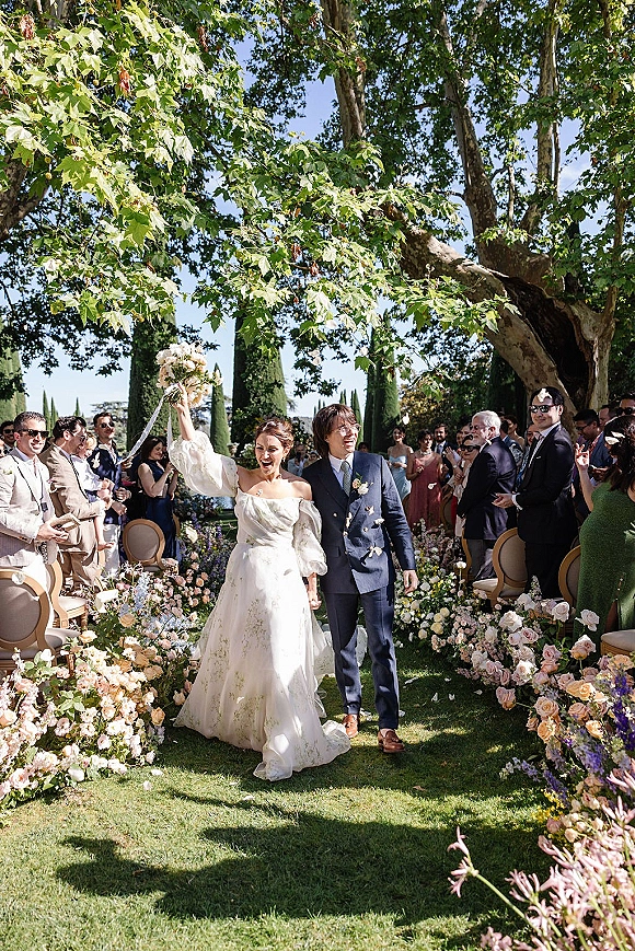 Wedding recessional as bride and groom walk the aisle, bride lifting bouquet with trailing ribbon, guests cheering in sunny garden setting