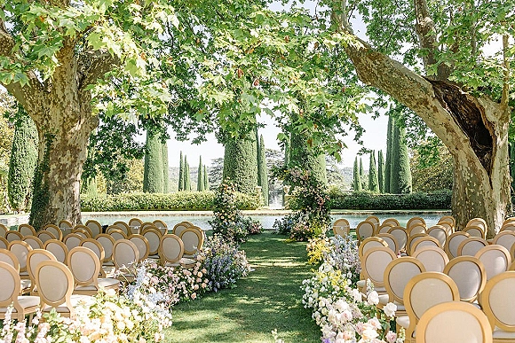 Outdoor ceremony setup with floral aisle decor leading to a greenery arch, symmetrical cream chairs on a lawn with fountain and tall trees