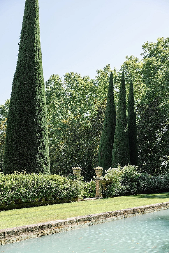 Garden scenery with manicured hedges, stone urn planters, and a stone wall beside a reflecting pool, framed by tall cypress trees under blue sky