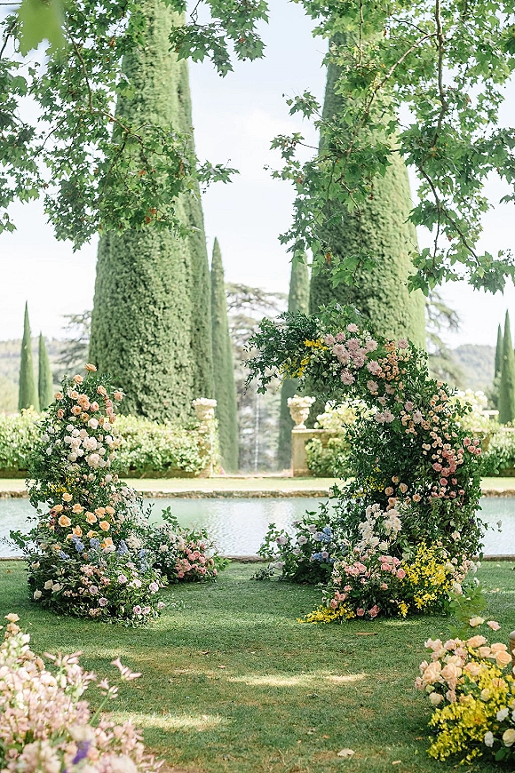 Wedding ceremony arch with an asymmetrical floral arch of garden roses and greenery by a reflecting pool and cypress trees