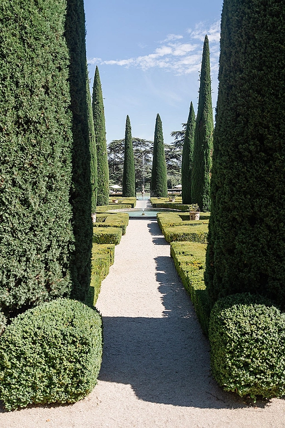 Garden walkway with a gravel path lined by trimmed hedges and cypress trees, leading to a reflecting pool and fountain under blue sky