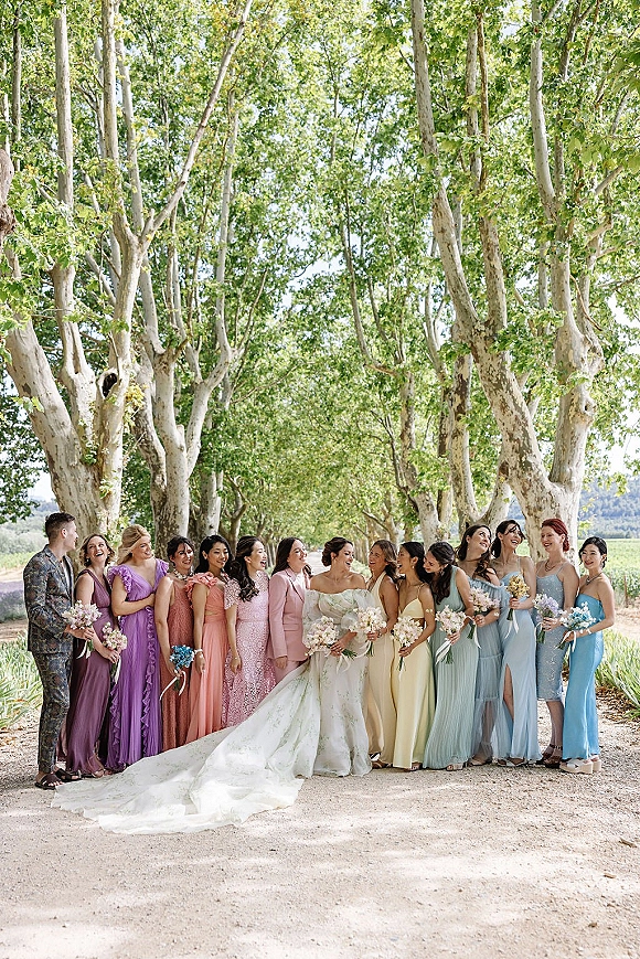 Wedding party portrait of bride with bridesmaids laughing in pastel dresses holding bouquets on a tree-lined path, long dress train flowing