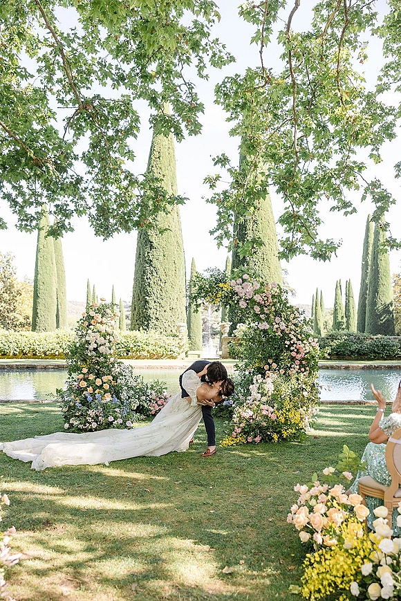Wedding kiss as the groom dips the bride in a long-train dress beneath a floral arch by a pool and fountain, guests seated on the lawn