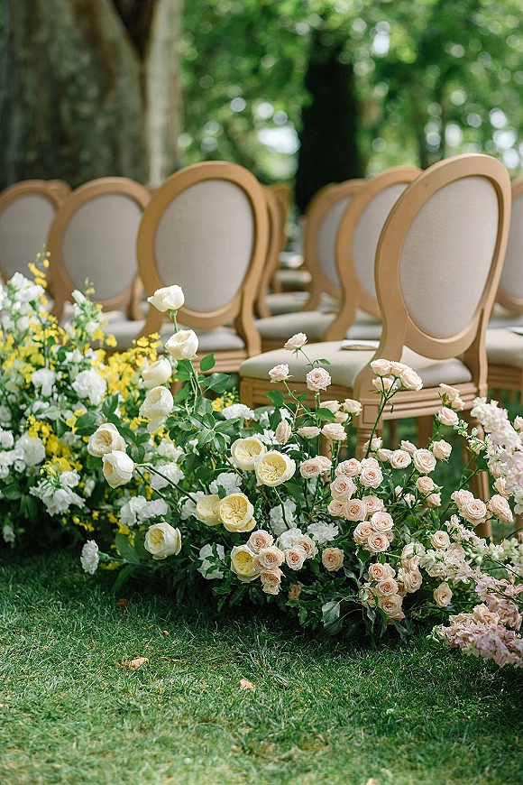 Ceremony aisle decor with garden roses lining the grass aisle beside oval-back and wooden chairs under trees and lush garden foliage