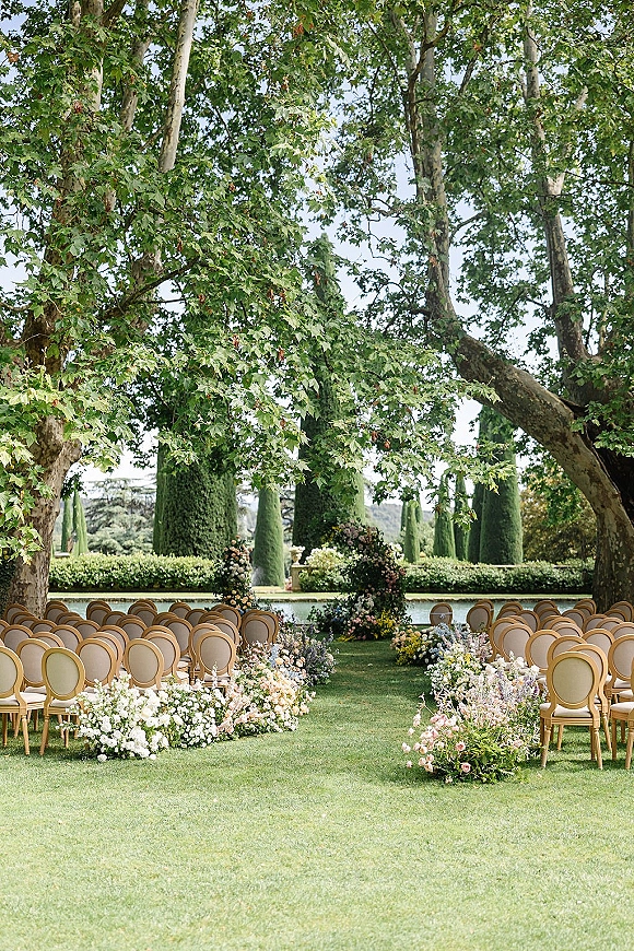 Ceremony setup for an outdoor wedding ceremony with round-back chairs and a floral arch beside a reflecting pool and hedges