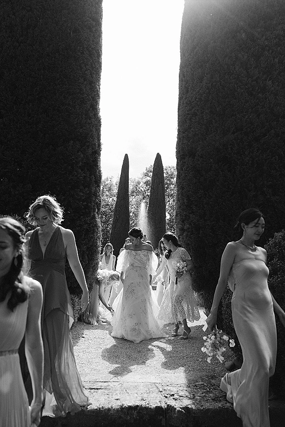 Wedding party portrait of bride with bridesmaids walking in a formal garden, holding bouquets in heels along a gravel path by a fountain