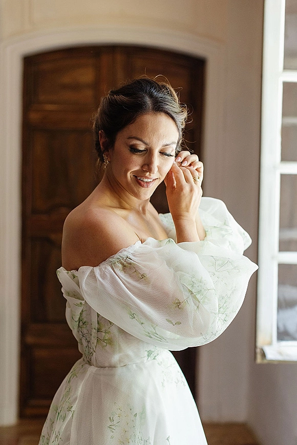 Bridal portrait of a bride getting ready, adjusting earrings in an off the shoulder floral dress by window light in a bedroom suite