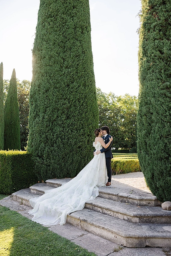 Wedding kiss portrait of bride and groom kiss on stone steps, her strapless gown with long train flowing in a formal garden of cypress trees