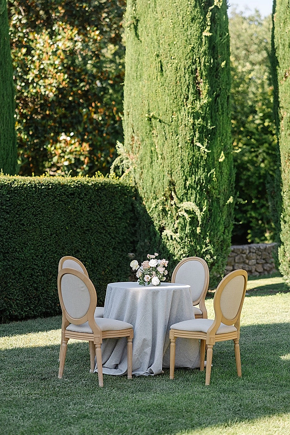 Reception tablescape with a round linen tablecloth and simple floral centerpiece, framed by upholstered chairs on a garden lawn by cypress trees.