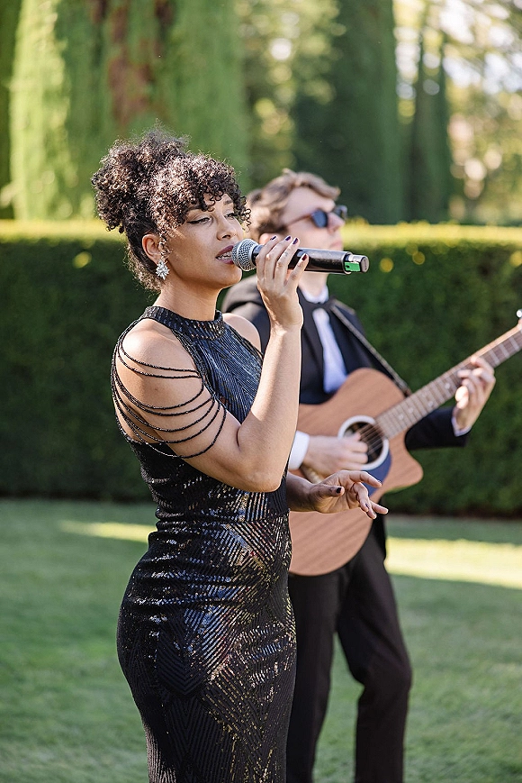 Wedding live music with a wedding singer in a black sequin dress singing into a handheld microphone as a guitarist plays on a garden lawn