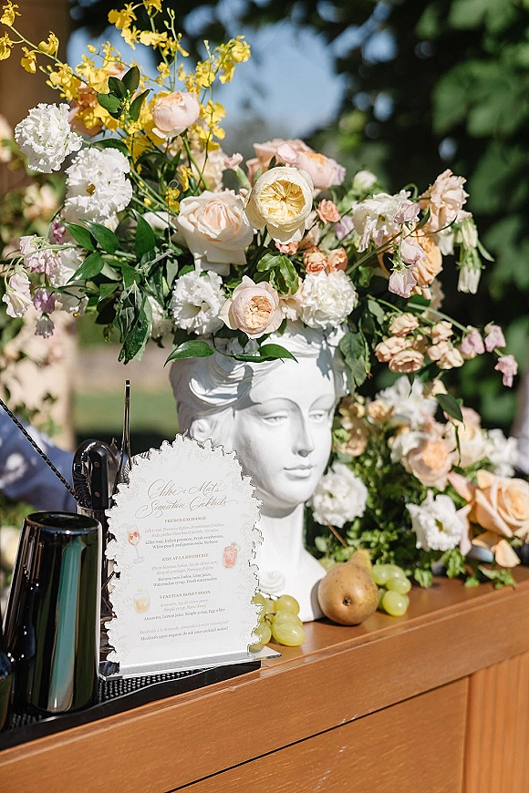 Wedding bar decor with a signature cocktail menu beside a classical bust vase of pastel roses, fruit and bar tools on a lawn in daylight