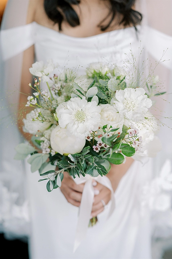 Bridal bouquet of white flowers and greenery tied with a ribbon, held against a bridal gown in soft natural light indoors