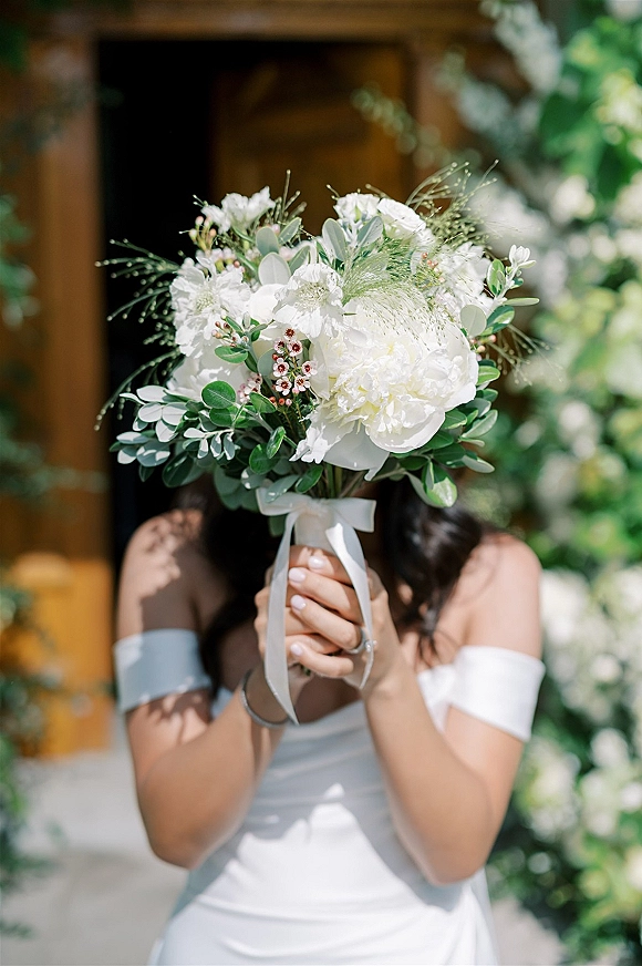 Bridal bouquet of white peonies with eucalyptus accents, ribbon wrap, and manicured hands with engagement ring by a leafy doorway backdrop