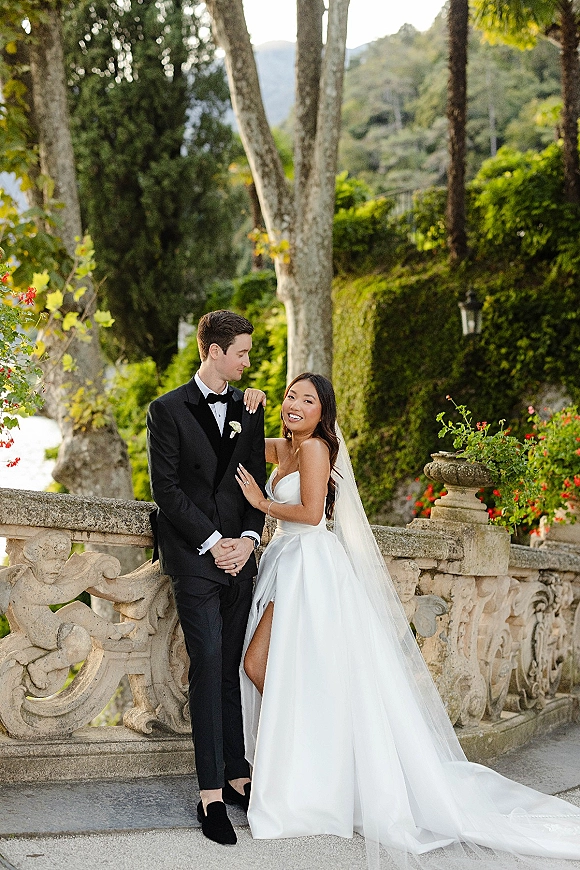 Couple portrait of bride in strapless wedding dress and veil leaning on groom in tuxedo by a stone balustrade with mountains behind